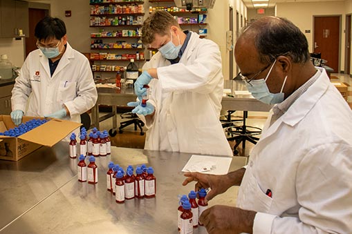 three men in white coats seal and pack bottles of hand sanitizer